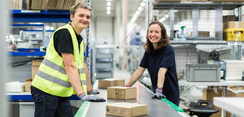 Smiling young man and woman standing by conveyor belt with boxes in distribution warehouse